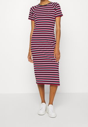 Vrouw die een midi-jurk met horizontale strepen in marineblauw, wit en rood draagt, met korte mouwen en witte sneakers.