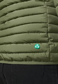 Veste en duvet vert olive présentant une texture matelassée horizontale, un matériau lisse et une étiquette en patch recyclé verte sur le bas à gauche.