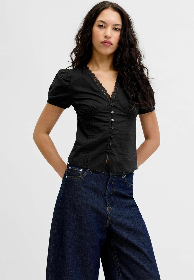 Femme aux cheveux longs et bouclés portant un chemisier noir à col en V boutonné avec un bord en dentelle et un jean large foncé, debout les mains derrière le dos.