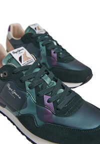 Zapatillas verdes con una mezcla de materiales lisos y texturizados, con acentos morados, cordones redondos y una etiqueta de marca en la lengüeta.