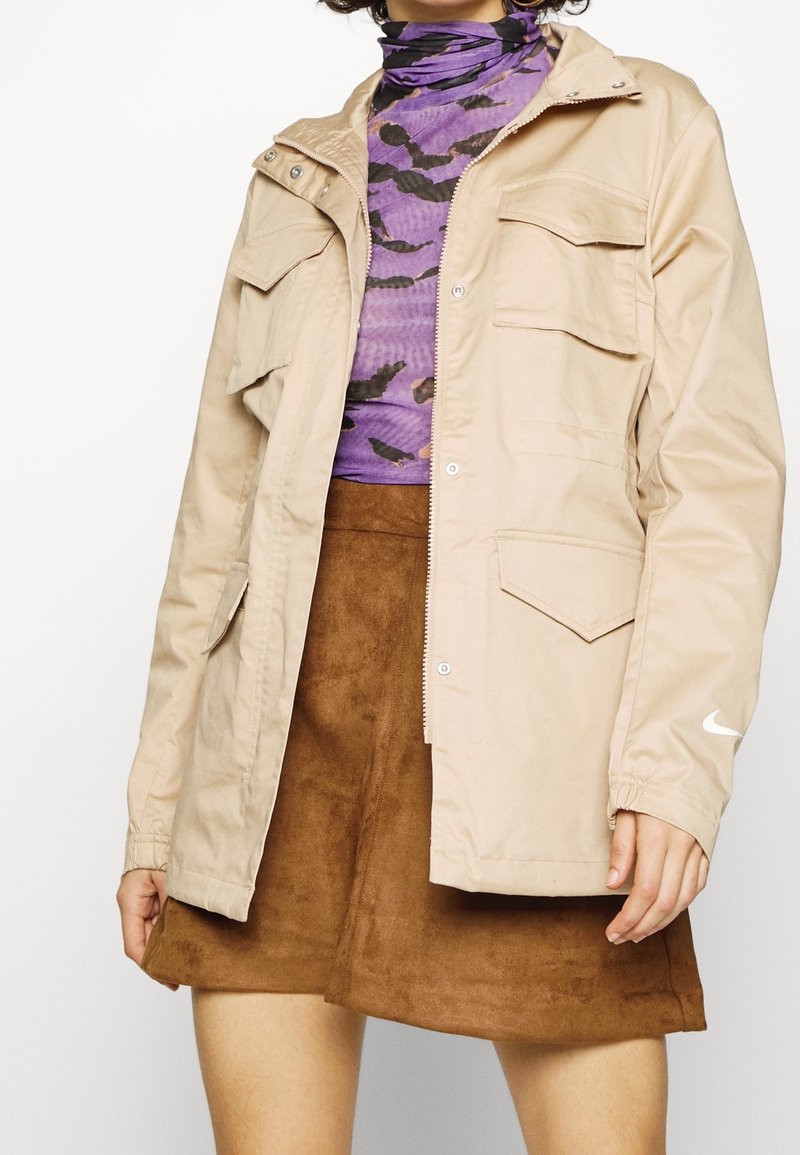 Beige nylon jacket with multiple front pockets, zip closure, and elastic cuffs. Paired with a purple patterned top and brown suede skirt.