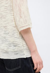 Manche de pull léger crème en maille lâche sur le bras, associée à un jean en denim bleu foncé avec des coutures visibles.