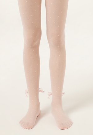 Beine, die durchsichtige rosa Strumpfhosen mit Punkten, verspielten Spitzen und Schleifen am Knöchel tragen, stehen auf einer weißen Oberfläche.
