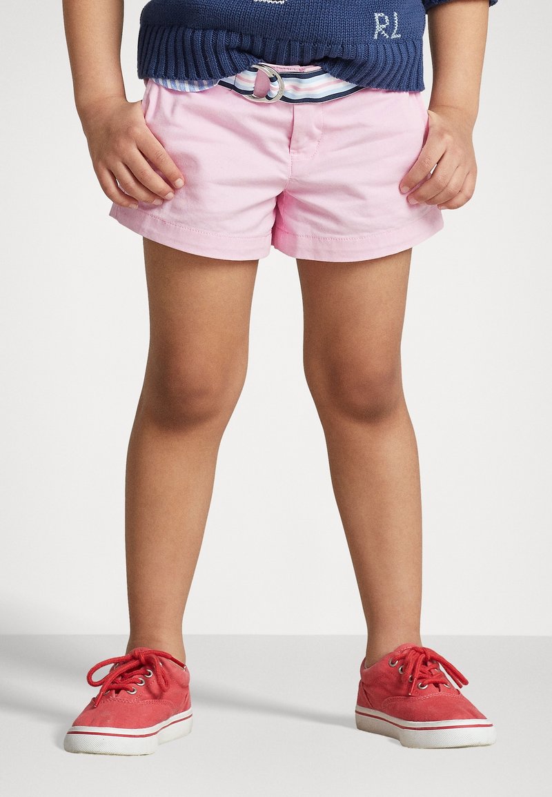 Rosa Baumwollshorts mit einem Gurtband, kombiniert mit roten Leinen-Sneakers mit weißen Gummisohlen. Schlichtes, lässiges Design.