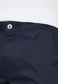Gros plan sur le devant d'un pantalon bleu marine avec un bouton en métal, une boucle de ceinture, et une chemise blanche rentrée à l'intérieur.
