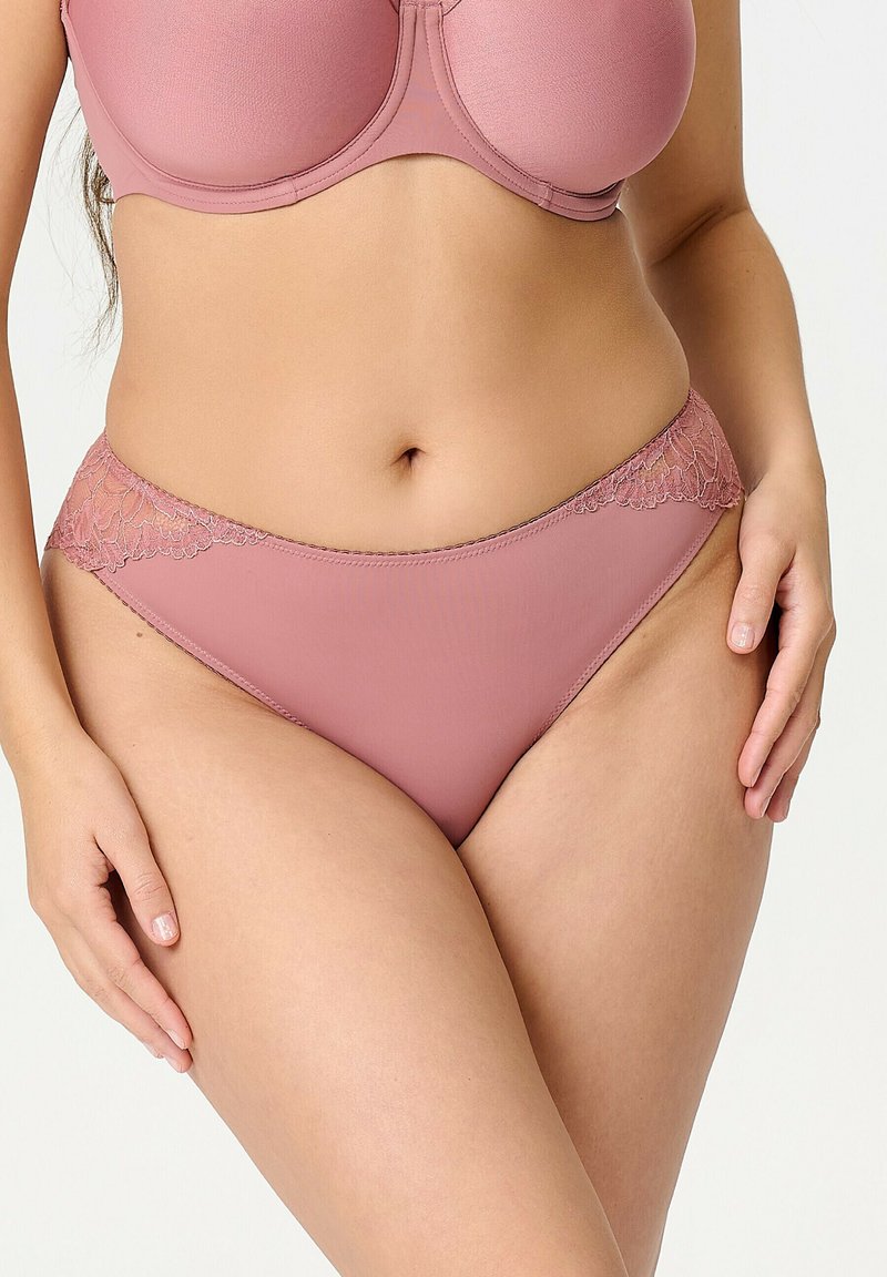 Femme portant un soutien-gorge rose poudré et une culotte assortie bordée de dentelle, debout avec les mains reposant sur les cuisses, sur un fond neutre.