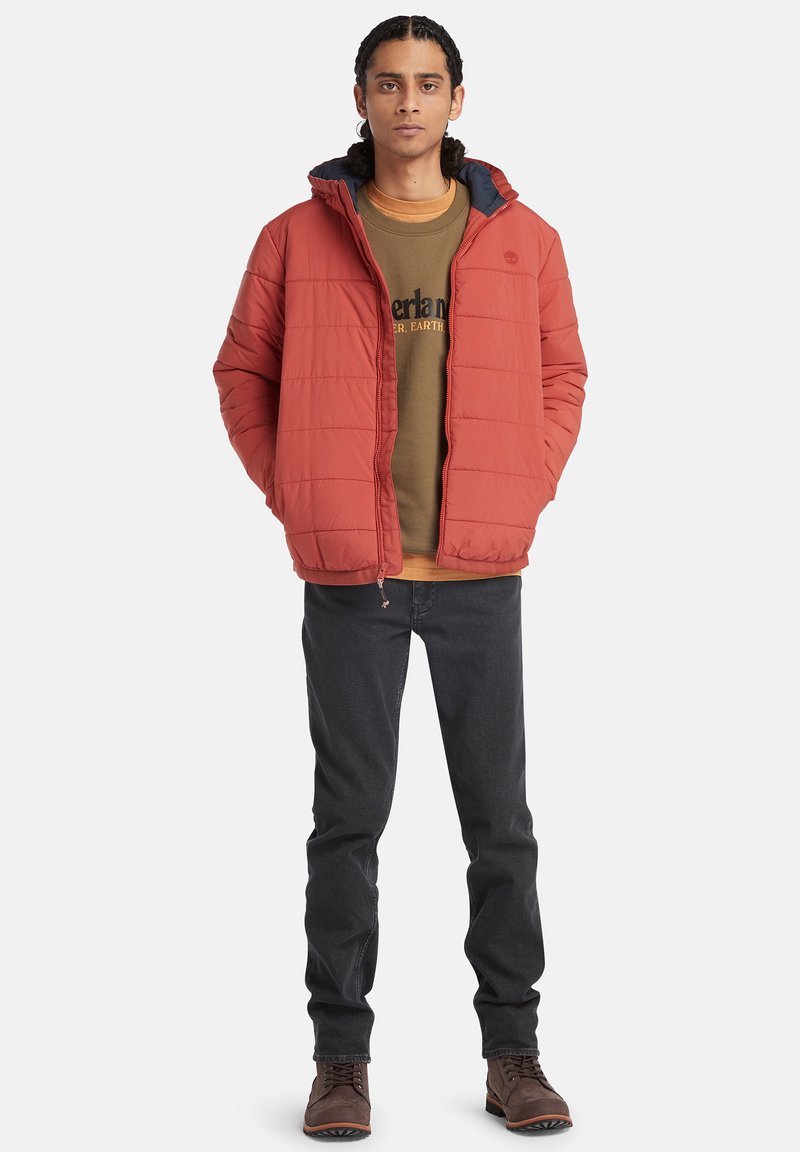 Rote gesteppte Jacke mit Reißverschluss und Kapuze, getragen über einem braunen Sweatshirt, kombiniert mit dunklen Jeans und braunen Stiefeln. Schlichte Gestaltung.