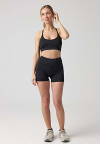 Top corto sportivo nero e pantaloni corti a vita alta realizzati in tessuto elasticizzato. Modella in piedi su uno sfondo neutro, indossando sneakers beige.