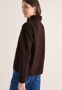 Femme portant une veste en polaire texturée marron foncé à col montant, associée à un jean en denim bleu, vue de profil et souriant légèrement.