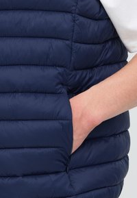 Ruka vklzávajúca do bočnej kapsy námornícky modrej prešívanej perovej vesty nosenej cez biele tričko s krátkym rukávom.