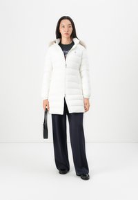 Hvid pufferjakke med lynlås fortil, hætte med faux fur-besætning og ribbede manchetter. Har lodret quiltet design. Bæres over navy brede bukser.