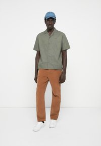 Short-sleeve, olive green button-up shirt with line pattern, paired with brown corduroy pants and white sneakers, topped with a light blue cap.