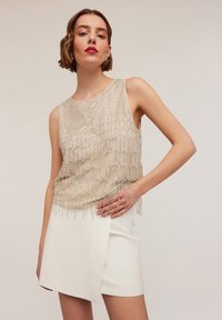 Beige top met kralen, voorzien van een textuurpatroon en franjes, gecombineerd met een witte wikkelrok. Glad materiaal en mouwloos ontwerp.