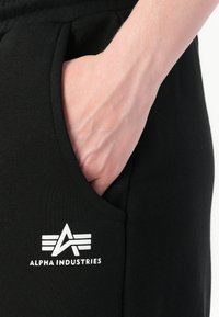 Hånd hviler i lommen på sorte bukser med hvidt Alpha Industries-logo under lommen.