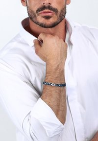 Blå geometrisk mönstrad armband gjort av metall, med en blandning av platta och upphöjda mönster, bärs på ett bar handled mot en vit skjorta.