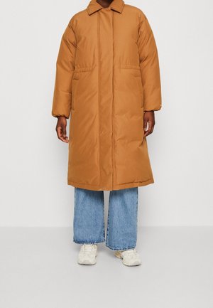 Osoba ubrana w długi, pikowany płaszcz w kolorze wielbłądzim z bocznymi kieszeniami, jasnoniebieskie, szerokie spodnie typu dzwony i białe trampki na neutralnym tle.