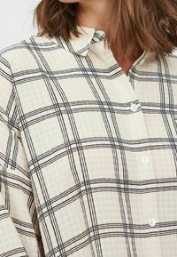 Woman wearing a beige button-up shirt with black checkered pattern, medium-length brown hair visible above the collar.