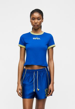 Kobieta nosząca niebieski krótki top Brasil z zielonym wykończeniem oraz pasujące niebieskie spodenki z żółtymi troczkami i logo Brasil na lewej nodze.