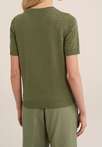 Back view of person wearing olive green textured short-sleeve sweater and matching loose pants against neutral background.