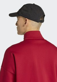 Červená košile s vysokým límcem a hladkým povrchem, kombinovaná s černou baseballovou čepicí s zakřiveným lemem a nastavitelným popruhem vzadu.