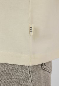 Étiquette en tissu beige marquée "BZB" attachée à l'ourlet d'une chemise de couleur crème portée sur un jean en denim gris.