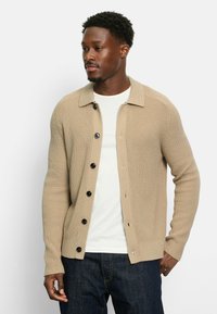 Beige Strickcardigan mit Kragen, sechs schwarzen Knöpfen, gerippter Textur und Seitentaschen, über einem weißen T-Shirt getragen.