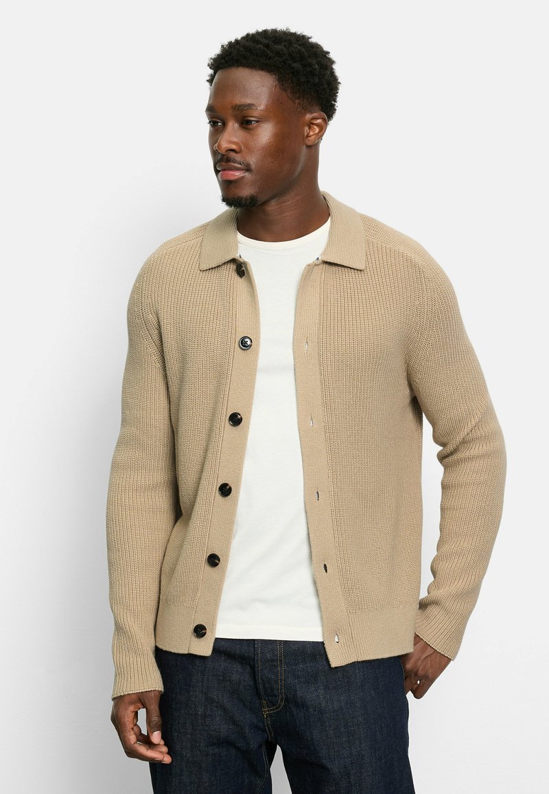 Beige Strickcardigan mit Kragen, sechs schwarzen Knöpfen, gerippter Textur und Seitentaschen, über einem weißen T-Shirt getragen.