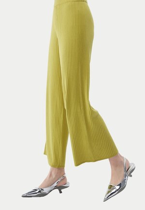 Personne portant un pantalon large côtelé vert citron et des escarpins argentés brillants à talons kitten et bride arrière sur fond clair.