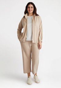 Beige sportjacka med huva, dragkedjestängning och sidofickor; kombinerad med matchande lösa byxor och vita sneakers.