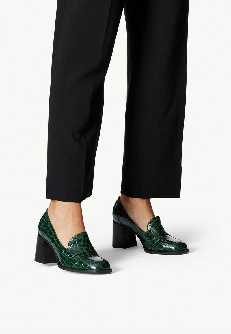 Femme portant un pantalon noir court et des mocassins à talons blocs texturés couleur crocodile vert foncé, debout sur un fond blanc.
