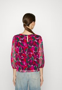 Blouse transparente multicolore avec un motif floral en rose, rouge et vert, dotée de manches bouffantes et d'une ouverture en trou de serrure à l'arrière.