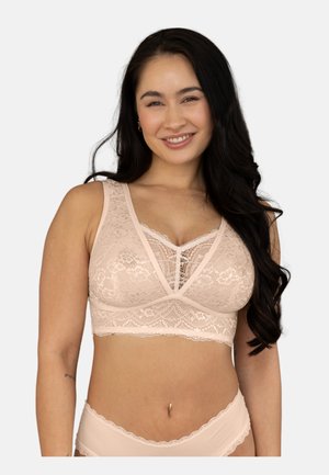 Smiling woman with long dark hair wearing a light pink lace bralette and matching underwear against a plain background.