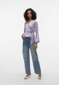 Blouse lilas à manches longues avec un lien à la taille, associée à un jean large bleu clair et des mules blanches.