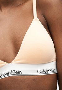 Γυναίκα που φοράει μπεζ τρίγωνο σουτιέν με λευκή ελαστική ταινία κάτω από το στήθος με το λογότυπο Calvin Klein.
