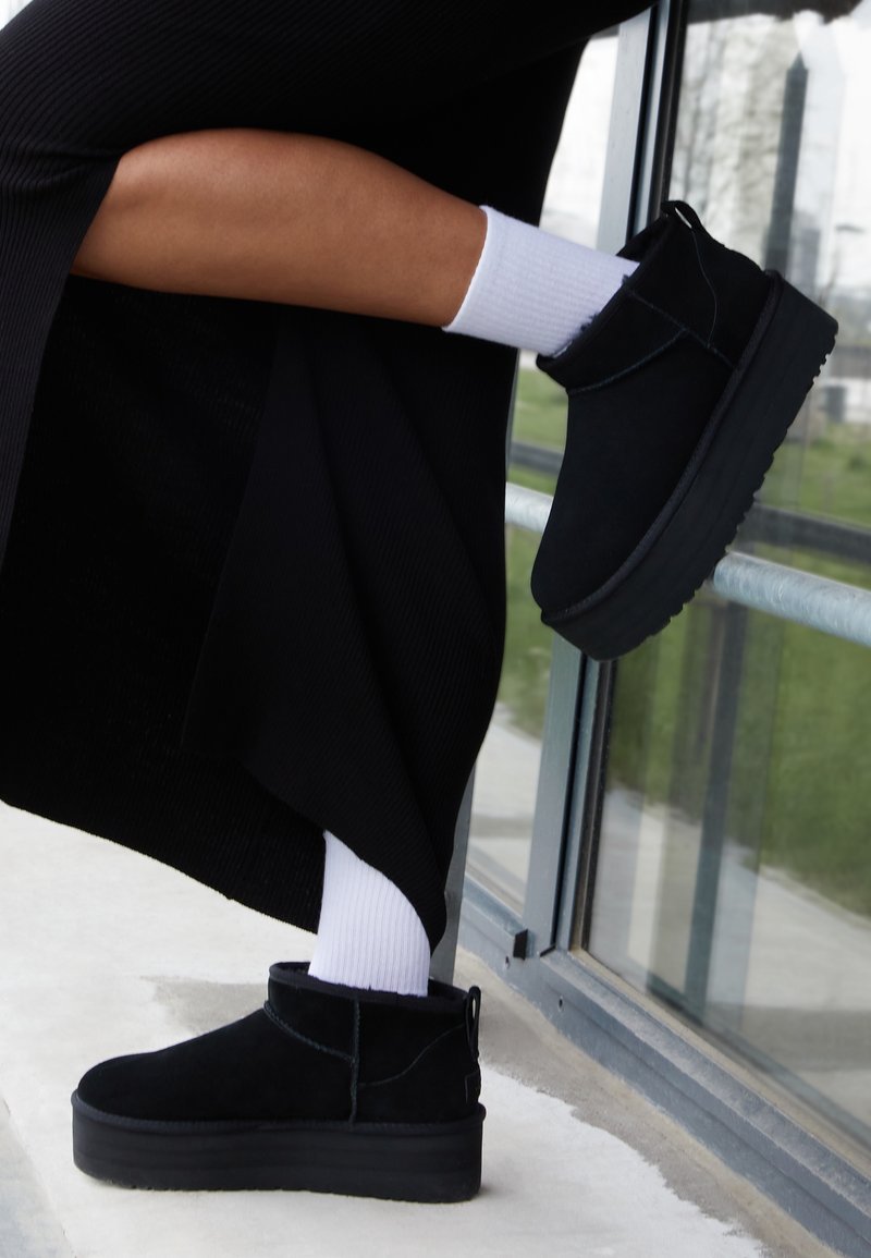 Jambes portant des chaussettes blanches et des bottines à plateforme noires, un pied levé contre une fenêtre en verre, vêtement noir visible.