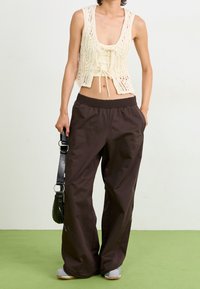 Bílý háčkovaný vest s krajkovým vzorem a zavazováním, v kombinaci s volnými hnědými kalhotami a botami se šedou podrážkou, držený černou taškou přes rameno.