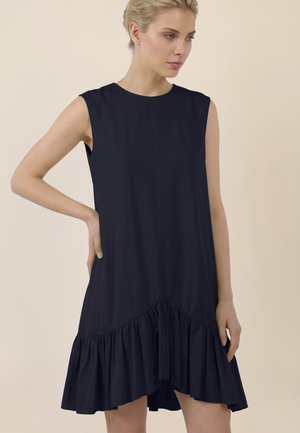 Frau mit blondem Haar, die ein ärmelloses schwarzes Kleid mit gerafftem Saum trägt, steht vor einem schlichten beigen Hintergrund, eine Hand in der Hüfte.