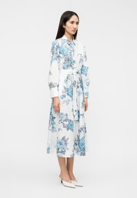 Λευκό φόρεμα με λουλουδάτο μοτίβο που περιλαμβάνει μπλε και γκρι τριαντάφυλλα, μακριά μανίκια και ζώνη στη μέση. Συνδυάζεται με λευκά παπούτσια μυτερά. Λεία υφή υφάσματος.