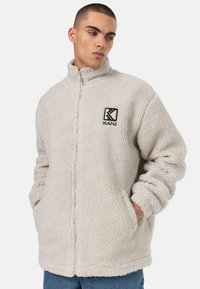 Heledbeez fleece-jakk, millel on ees lukuga, kõrge kraega ja küljetaskud. Rinnal on must "KANI" logo. Pehme, tekstureeritud kangas.
