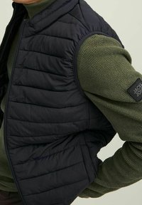 Mustan pehmustetun liivin, jossa on tikkikuvio, päällä tekstiilistä valmistettu oliivivihreä neule. Varustettu vetoketjulla ja olkapäällä logopatch.