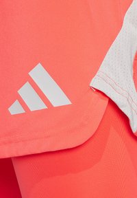 Koraal sportshirt met een wit logo met drie strepen, voorzien van textuur op de grijze mouwen en een gladde stof. De zoomlijn is licht afgerond.
