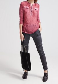 Rode, lange mouwen, knoopsluiting shirt met twee borstzakken; gecombineerd met grijze, strakke broek en een zwarte suède handtas met kettingaccenten.