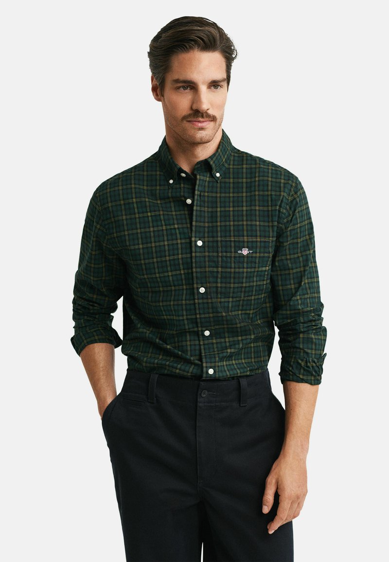 Camicia a quadri verde e blu navy con colletto a risvolto, dotata di maniche lunghe e un piccolo logo sul petto. Pantaloni neri.