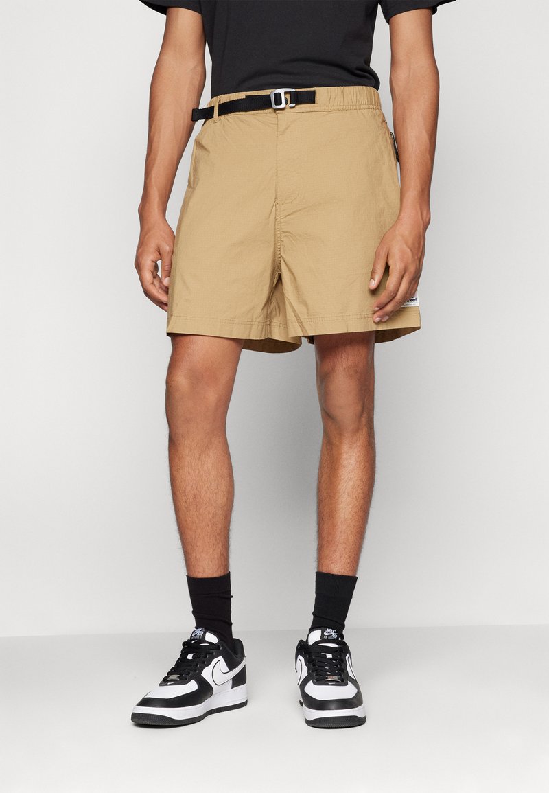 Lysbrune knælange shorts med en struktureret finish, der har et sort bælte og sidelomme. Særskilt med sort og hvidt sneakers.