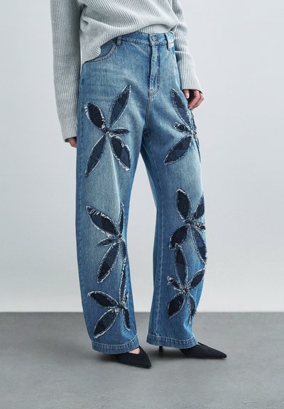 Jeans en denim bleu clair avec de grandes pièces florales bleu foncé, présentant des bords effilochés. Taille haute, coupe décontractée et design à jambes larges.