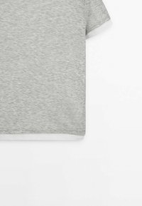 T-shirt gris à manches courtes superposé à une chemise blanche, montré de la taille à la poitrine sur un fond blanc.