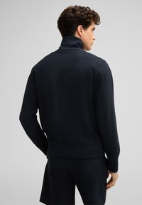 Dunkelblaues Zip-Jackett mit strukturiertem Muster, hohem Kragen sowie elastischen Bündchen und Saum, getragen mit passenden Shorts.