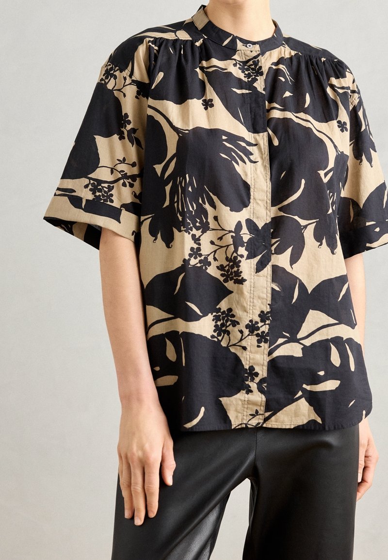 Personne portant une chemise beige à manches courtes avec de grands motifs floraux noirs et un pantalon noir, debout devant un fond clair uni.