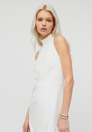 Blonde woman with loose hair wears sleeveless white high-neck dress and silver bracelets against plain light background.