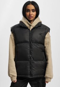 Schwarze wärmende Weste ohne Ärmel mit Reißverschluss, zwei Seitentaschen und glänzendem Finish, darüber getragen einen beigen Hoodie.